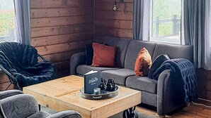 Ein gemütlicher Raum mit einer Holzwand, einem grauen Sofa mit roten und schwarzen Kissen, einem schwarzen Stuhl und einem Holztisch mit einem Buch und Teekannen.