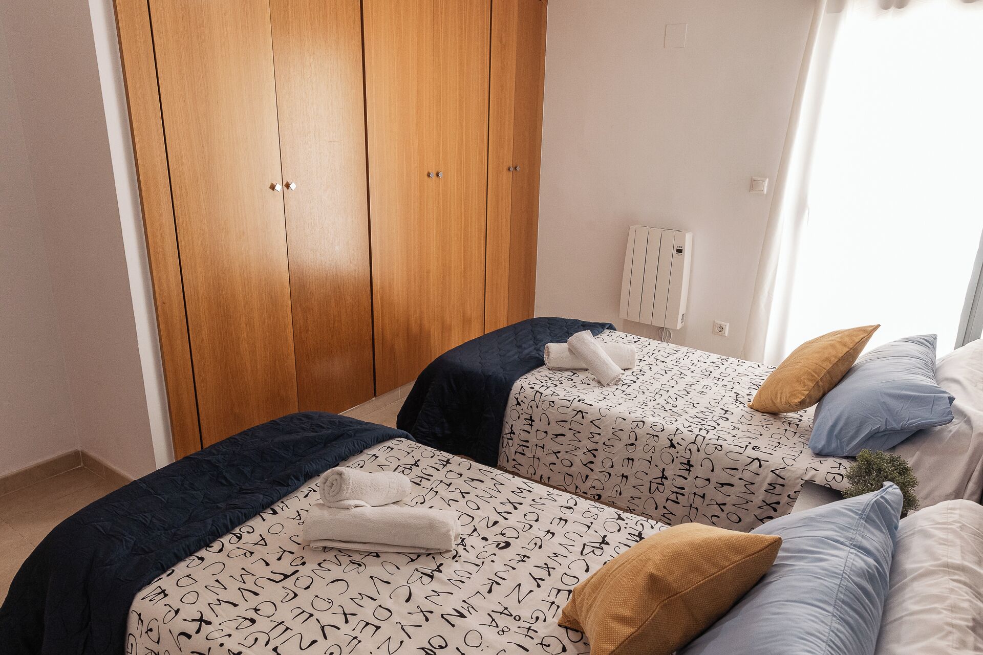 Un dormitorio con dos camas, cada una con una colcha y almohadas estampadas, un armario de madera y una pared blanca con un ventanal pequeño.