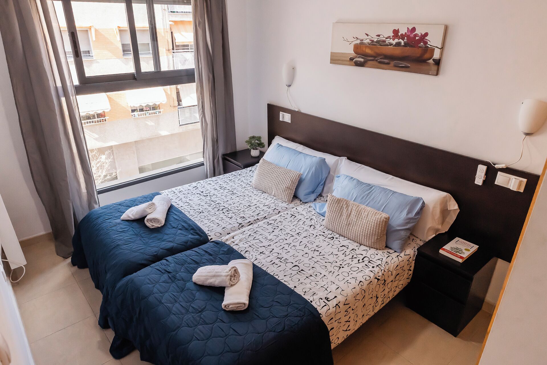 A double bed with a patterned bedspread, two pillows, and folded towels. There is a small bedside table with a lamp and a power outlet. A painting hangs on the wall above the bed.