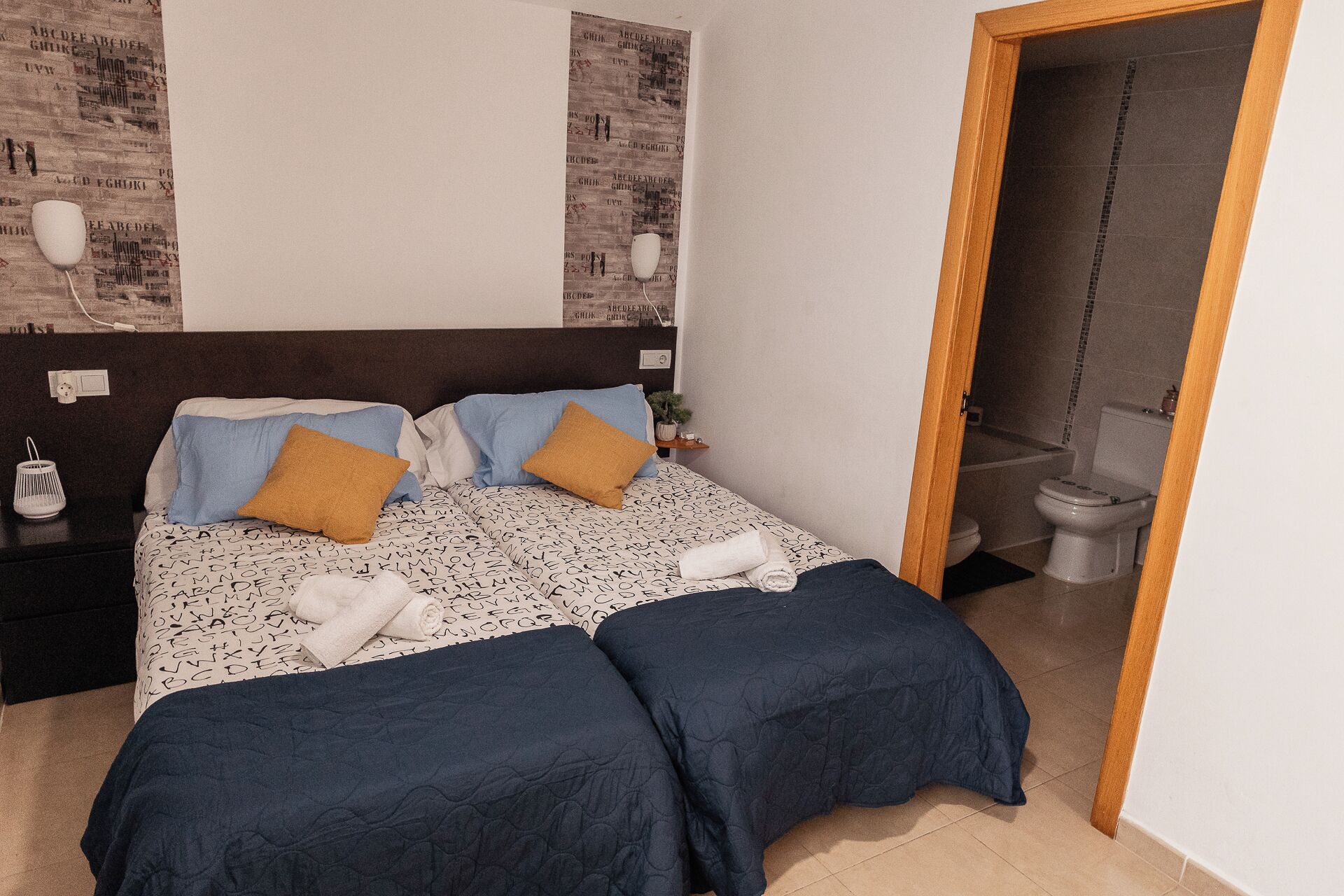 A bedroom with a bed, two bedside tables, a nightstand, a wall-mounted lamp, and a bathroom visible through an open door.