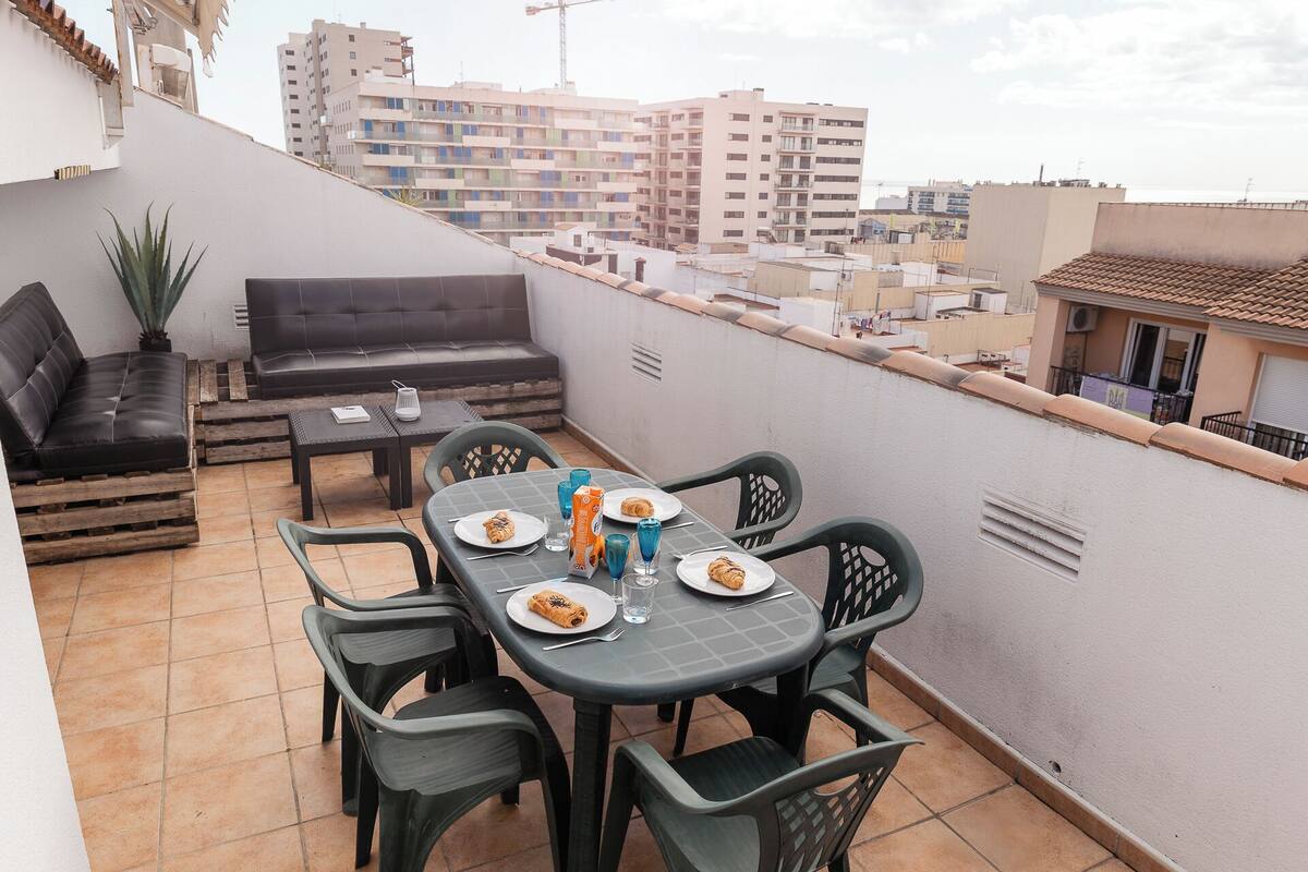 Une terrasse sur le toit, avec une table à manger pour deux, entourée de chaises vertes. On y trouve un canapé noir et une petite table avec une plante. La terrasse offre une vue sur le paysage urbain, avec des bâtiments en arrière-plan.