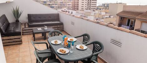 Una terraza en la azotea con una mesa de comedor para dos personas, rodeada de sillas verdes. Hay un sofá negro y una mesita con una planta. La terraza tiene vistas a un paisaje urbano con edificios al fondo.