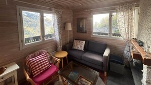 A cozy cabin interior with a wooden ceiling, two windows with curtains, a sofa, a chair, a small table, and a lamp. - Chalet With Garden and Mountain View (Vex)
