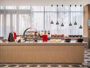 Zona de recepción de un hotel con una barra de buffet, un amplio ventanal con cortinas translúcidas y un diseño moderno.