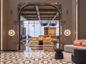 Un hall d’hôtel moderne avec un sol à motifs géométriques, un espace salon central doté de canapés et d’une table basse, et une grande porte en arc donnant sur une salle à manger avec un bar.