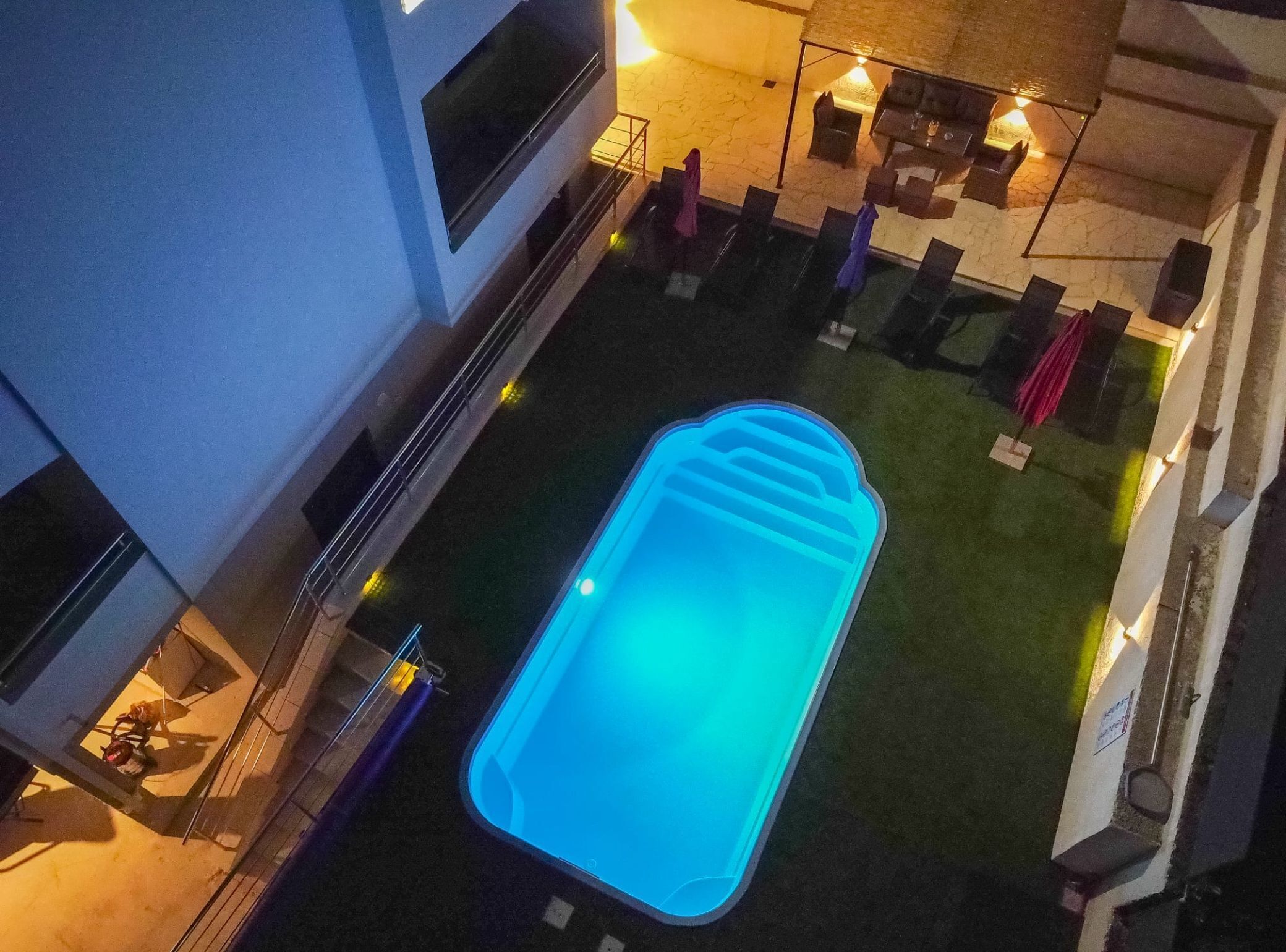 Vue aérienne d’un espace piscine, avec une piscine illuminée de lumière bleue, entourée d’une petite pelouse et de quelques chaises longues.