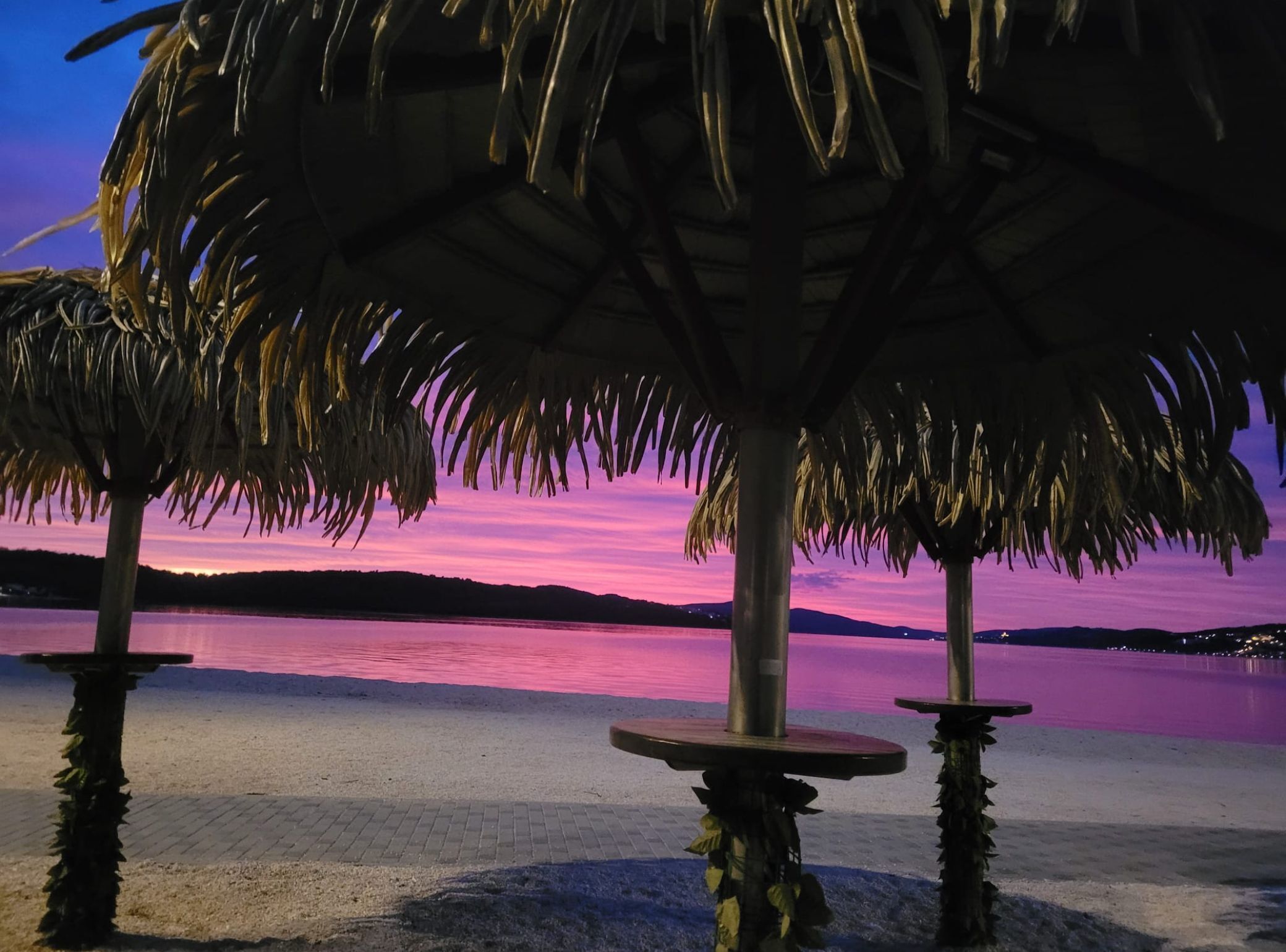 A beachside view with palm trees and a sunset over water.
