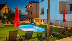 A pool area with lounge chairs and umbrellas, surrounded by a well-maintained lawn and a building with a balcony. - Apartments Neli (Okrug)