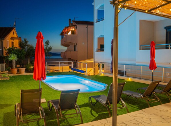 A pool area with lounge chairs and umbrellas, surrounded by a well-maintained lawn and a building with a balcony. - Apartments Neli (Okrug)
