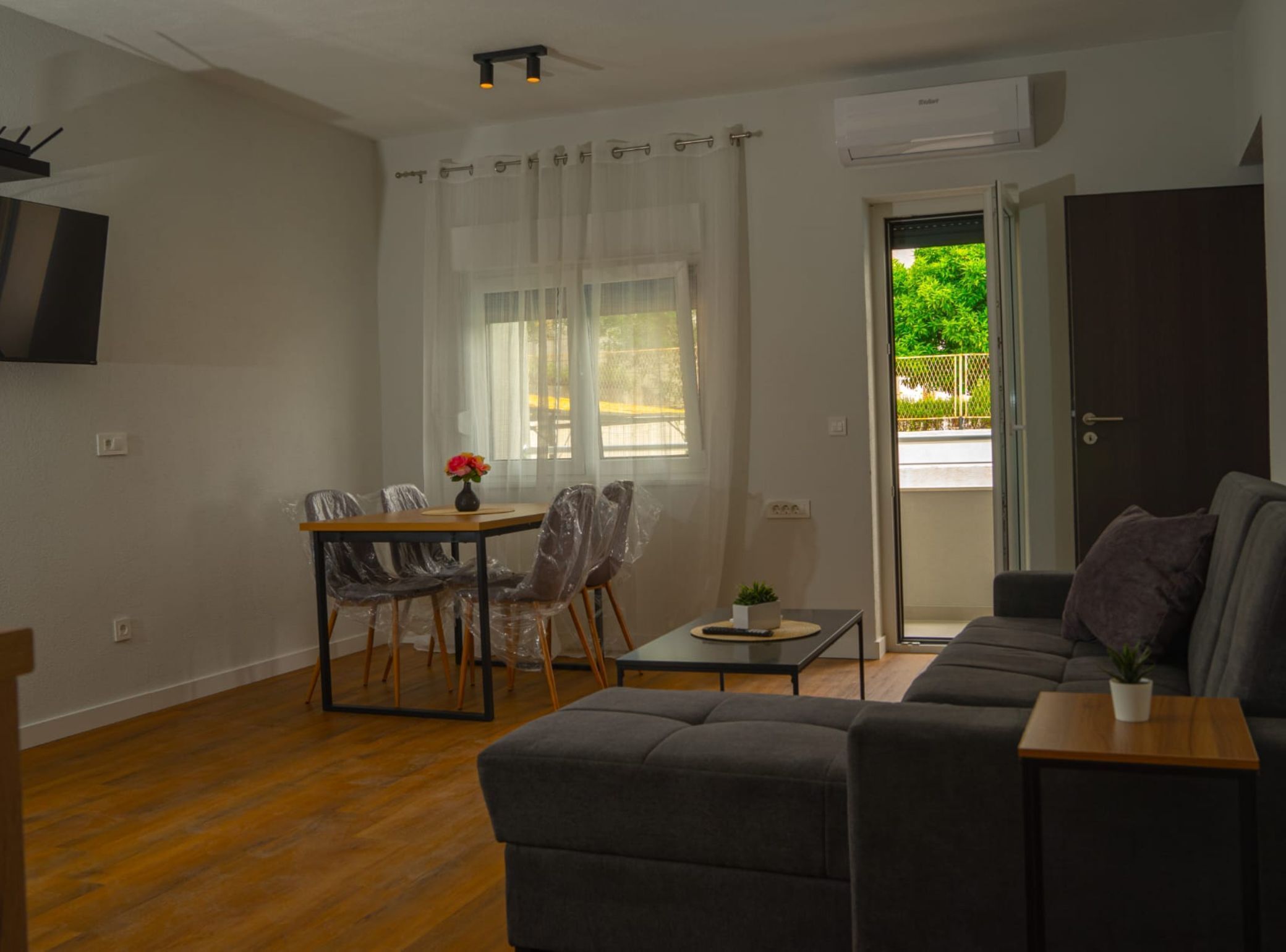 A living room with a sofa, a dining table with chairs, a small table with a plant, and a sliding glass door leading to an outdoor area.