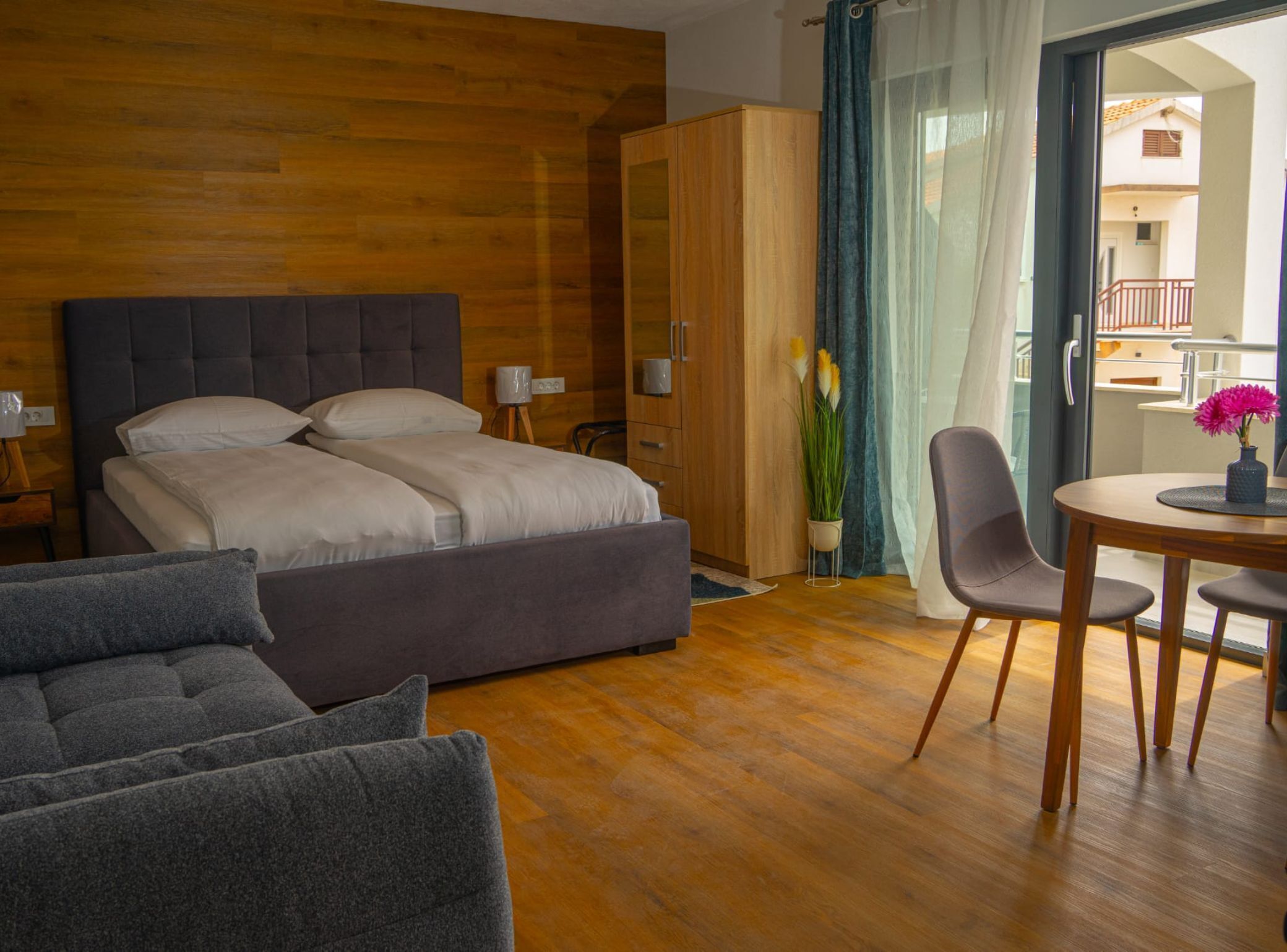 A modern bedroom with a large bed, a wooden wardrobe, a dining area with a table and chairs, and a balcony with a view.
