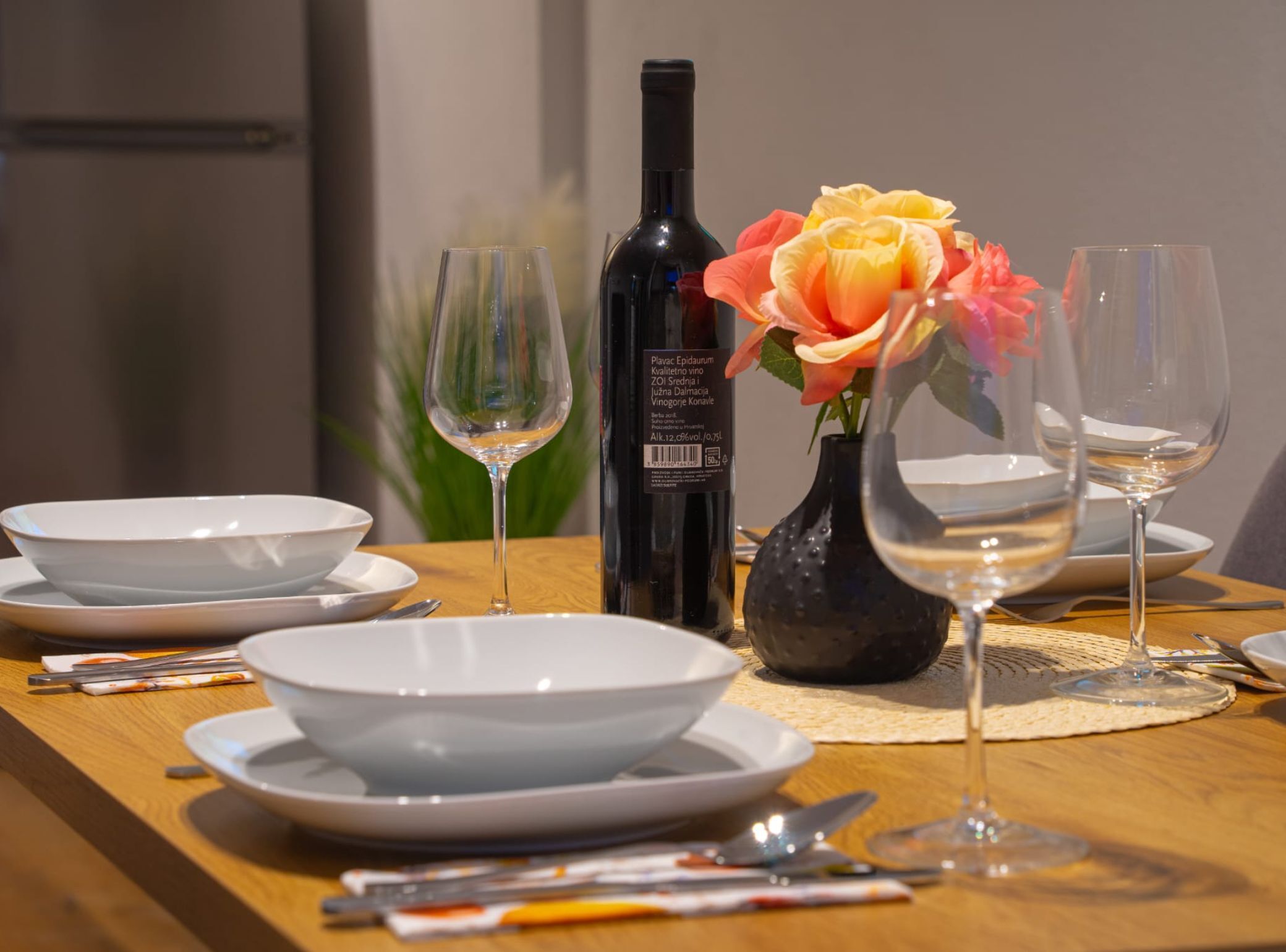 A dining table with white plates, wine glasses, and a bottle of wine. A vase with a yellow flower is also visible.