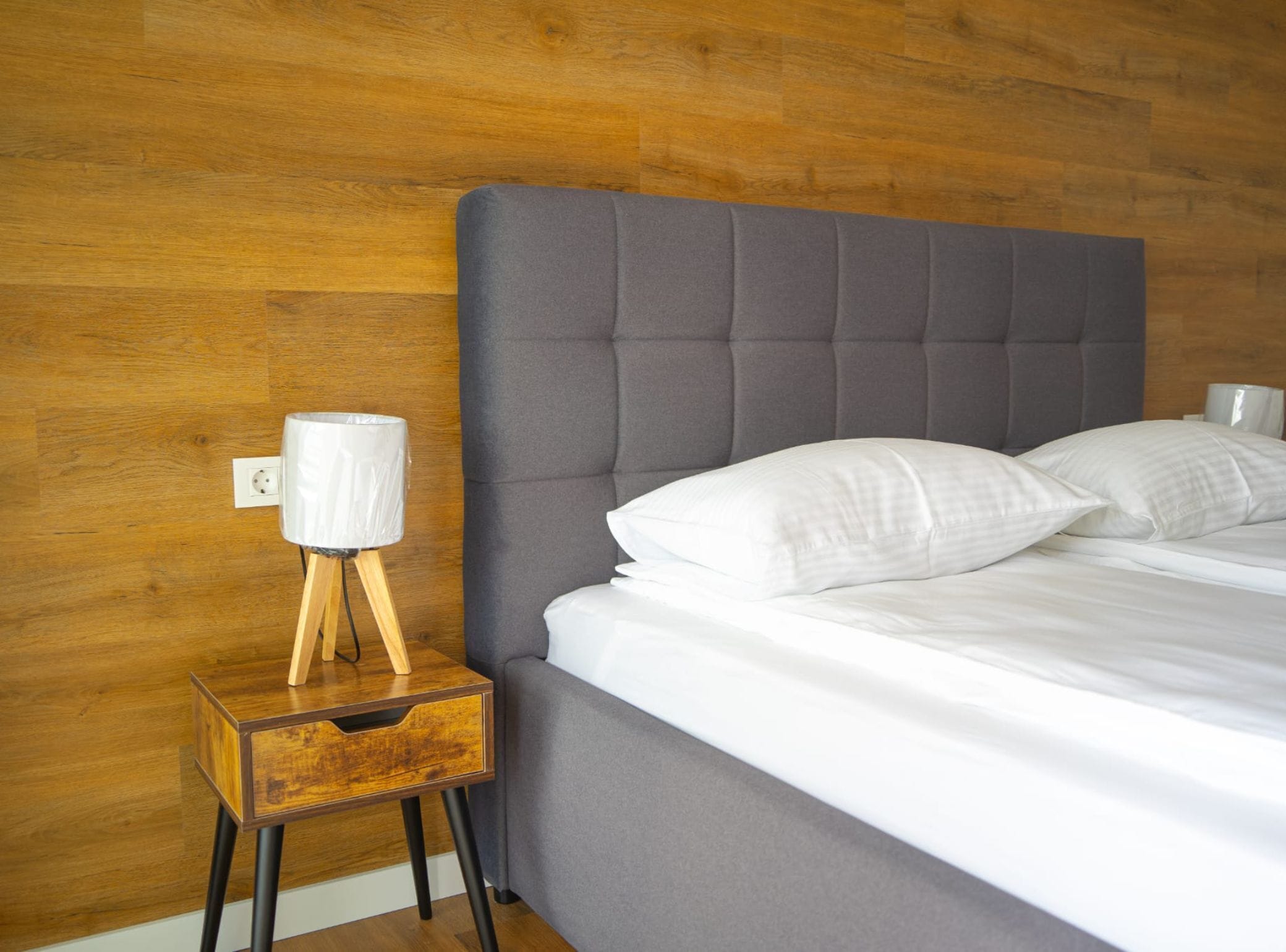 Un letto rifatto con lenzuola e cuscini bianchi, una testiera grigia imbottita e un piccolo comodino in legno con una lampada.
