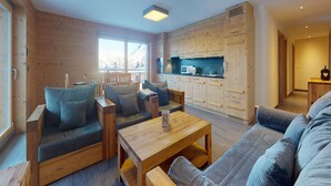 A modern living room with wooden furniture, a kitchen area, and a large window providing natural light.