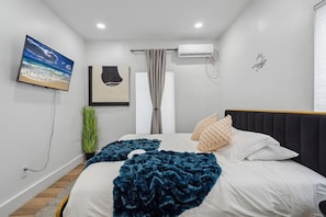 Un dormitorio con cama, aire acondicionado de pared, ventana con cortinas, una planta y obras de arte.