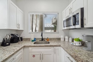 A kitchen with white cabinets, a granite countertop, a stainless steel sink, a window with blinds, and a microwave.