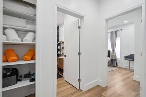 A hotel room with a walk-in closet, a bed with a grey headboard, a small desk, and a window with curtains.