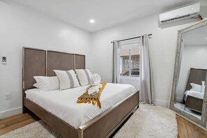 Una habitación de hotel moderna con una cama grande, un cabecero de madera, una ventana con cortinas, un espejo y un equipo de aire acondicionado.