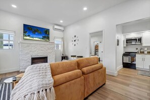 Una sala de estar moderna con chimenea de piedra, un sofá grande y una cocina con armarios y electrodomésticos blancos.