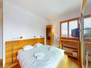 Een opgemaakt bed met witte lakens en handdoeken, een houten hoofdbord, een nachtkastje met een lamp, en een groot raam met uitzicht op zee.
