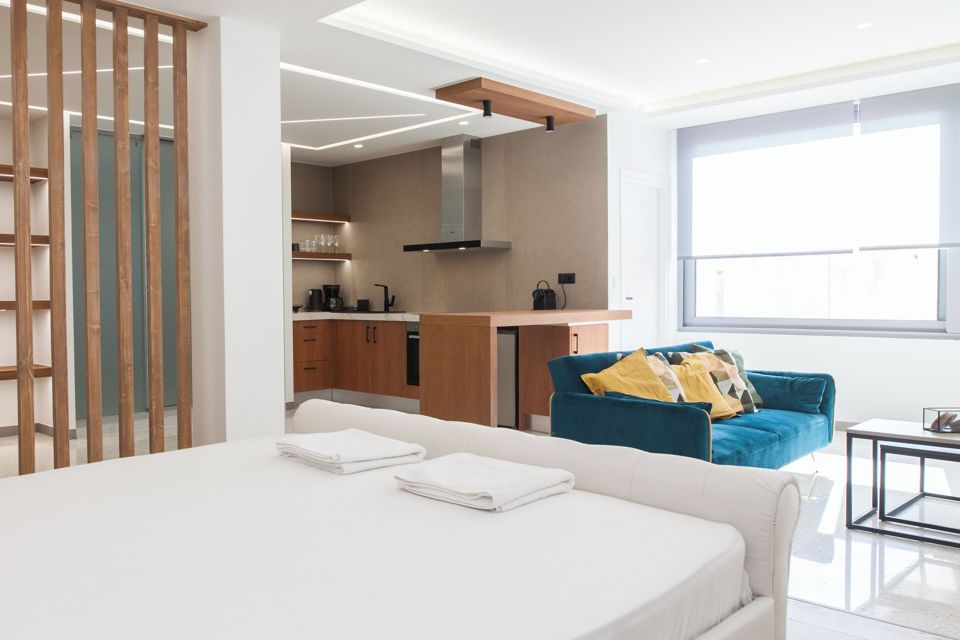 Une chambre d’hôtel moderne avec un grand lit, un canapé bleu et un coin cuisine visible à travers une porte ouverte.