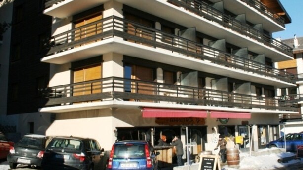 A multi-story building with balconies, a ground-level restaurant, and cars parked in front. - Pleasant Apartment With Balcony (Veysonnaz)