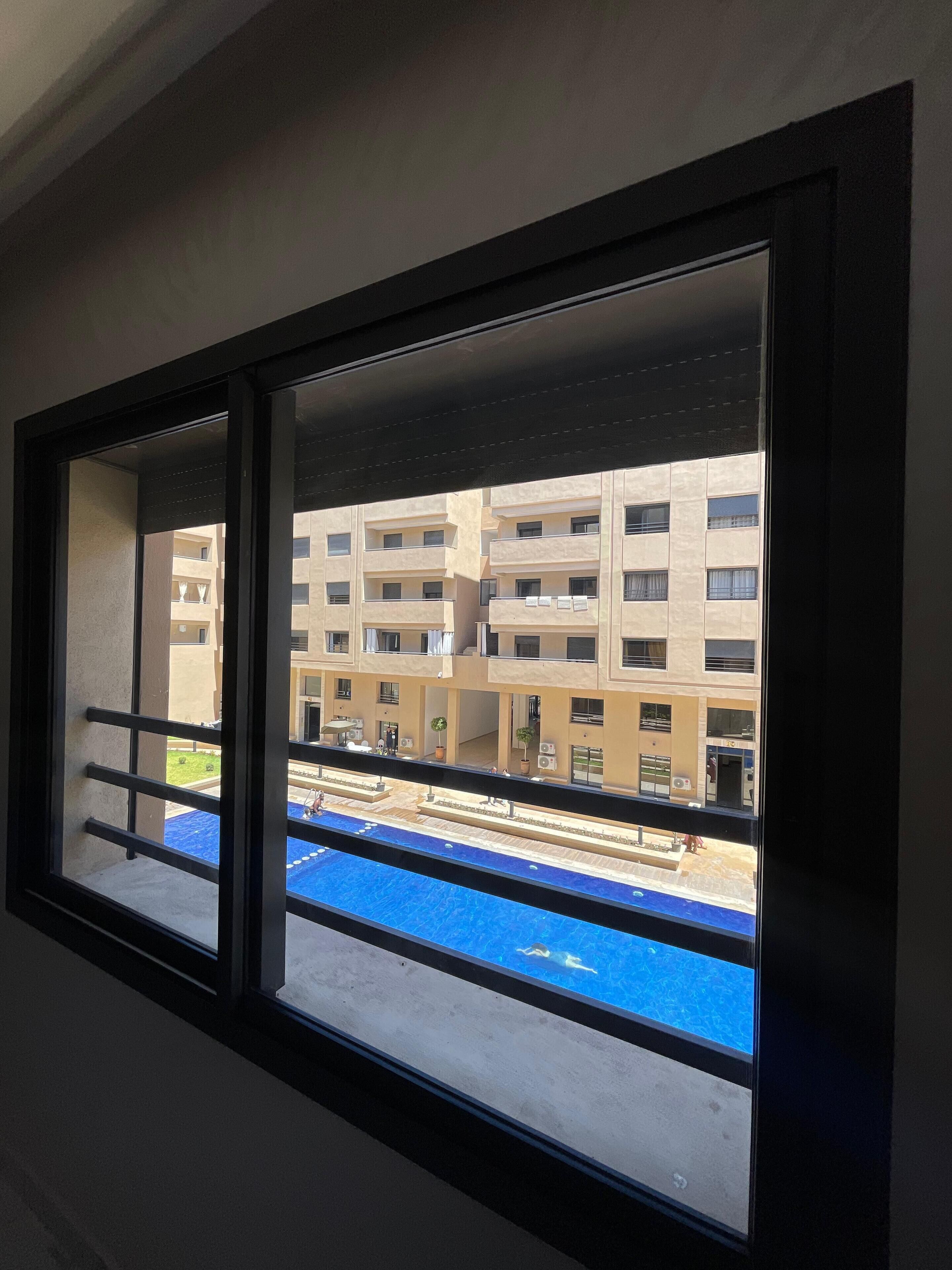 Une fenêtre donnant sur un immeuble doté de nombreux balcons et sur un espace piscine bleu.