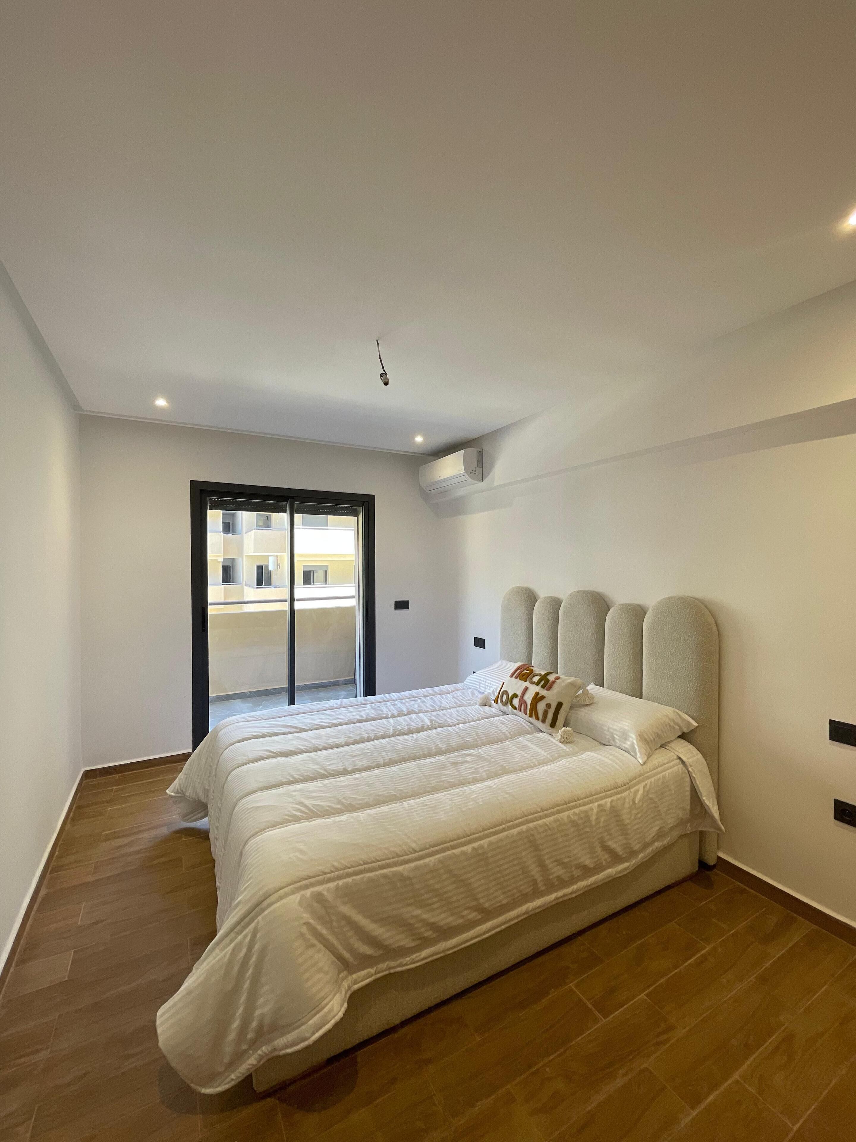 A modern bedroom with a large bed, a sliding glass door to a balcony, and a ceiling-mounted air conditioning unit.