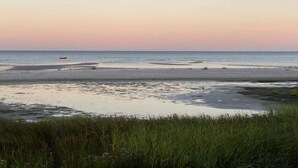 Une plage avec un bateau visible au loin et des dunes herbeuses au premier plan.