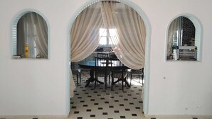A dining area with a table and chairs, a patterned floor, and arched windows with curtains. - VILAA TEJ EL MOULK (Somâa)