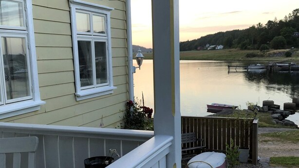 Ett hus med en veranda som vetter mot en lugn sjö, ett trädäck och en båt som ligger förtöjd vid bryggan.