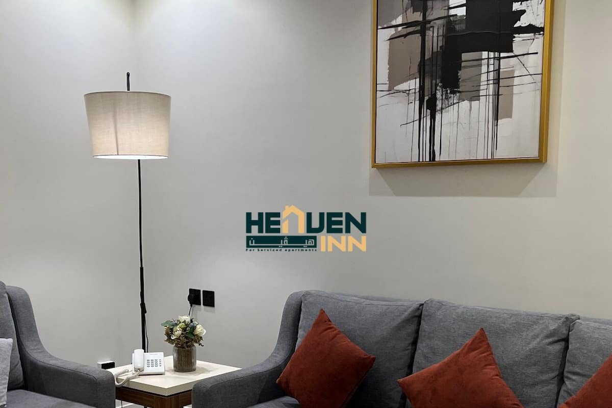 A modern living room with a grey sofa, a wooden coffee table, and a floor lamp. There is a framed abstract painting on the wall and a logo for 'HEVEN INN' on the wall.