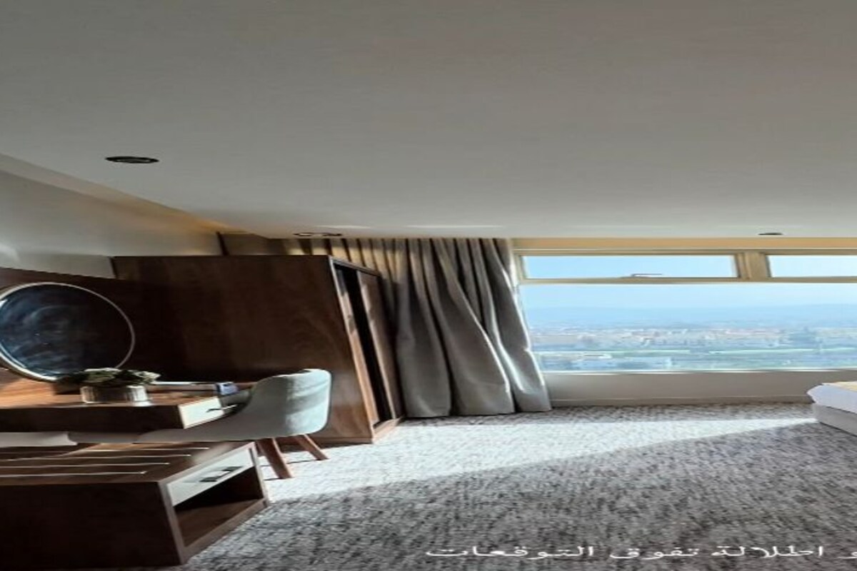 A hotel room with a large window, a desk with a chair, a bed, and a small table with a lamp.