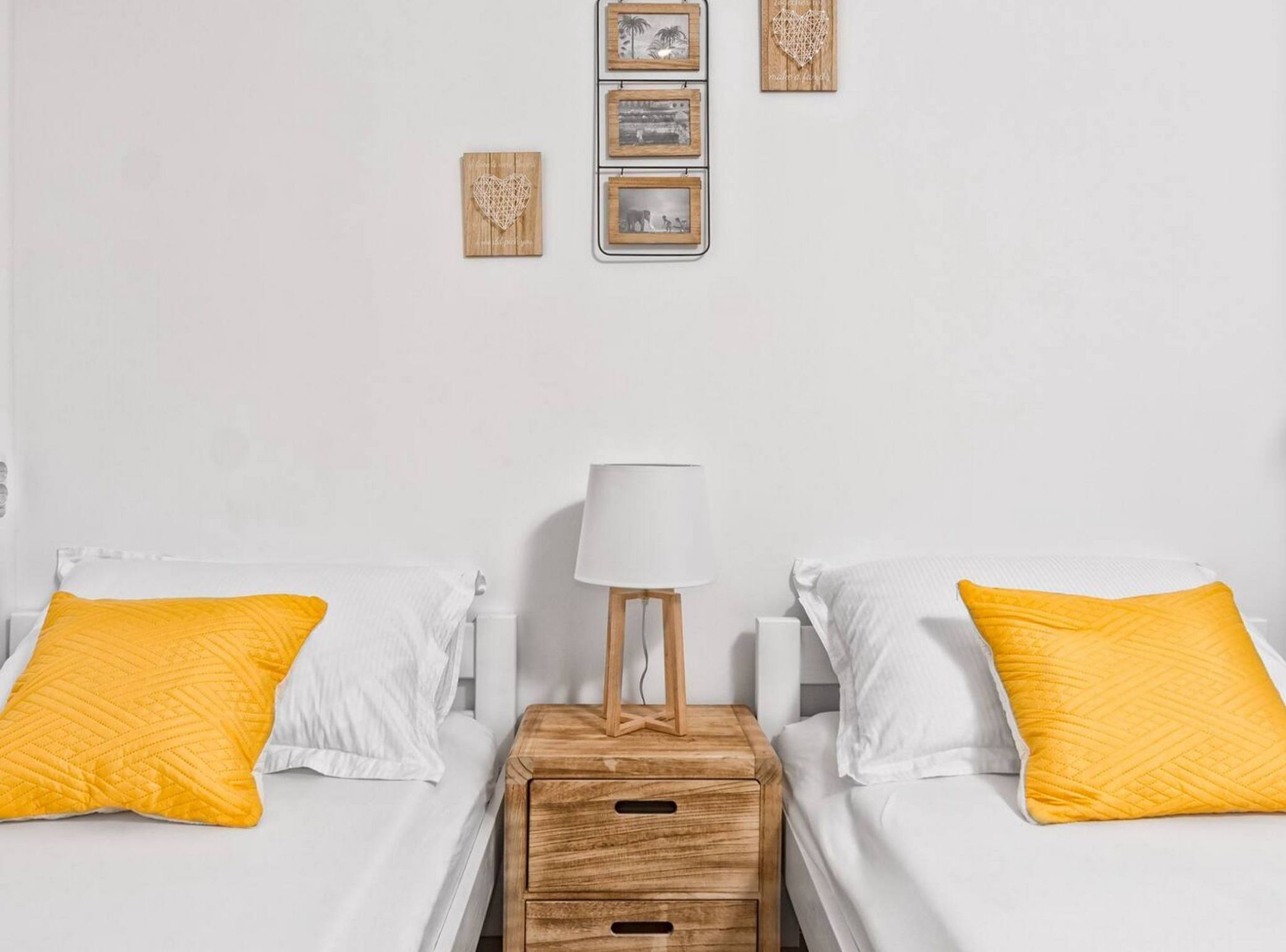 Habitación de hotel con dos camas, cada una con colchón blanco y almohada amarilla. Hay una mesita de noche de madera con una lámpara entre las camas.