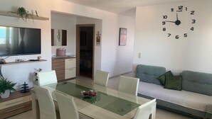 A modern living room with a dining table, chairs, a sofa, a TV, and a wall clock. - Mastrinka Bright Apartment With Free Parking and Modern Amenities (Trogir)