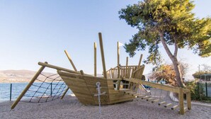 A wooden pirate ship play structure in a park with a clear view of the sea and hills in the background. - Mastrinka Bright Apartment With Free Parking and Modern Amenities (Trogir)