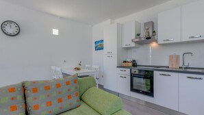 A modern kitchen with white cabinets, a built-in oven, and a sink. There is a dining area with a white table and chairs, and a green sofa with patterned cushions. A clock is mounted on the wall. - Seaside Getaway Holiday Home With Patio Slatine (Split)