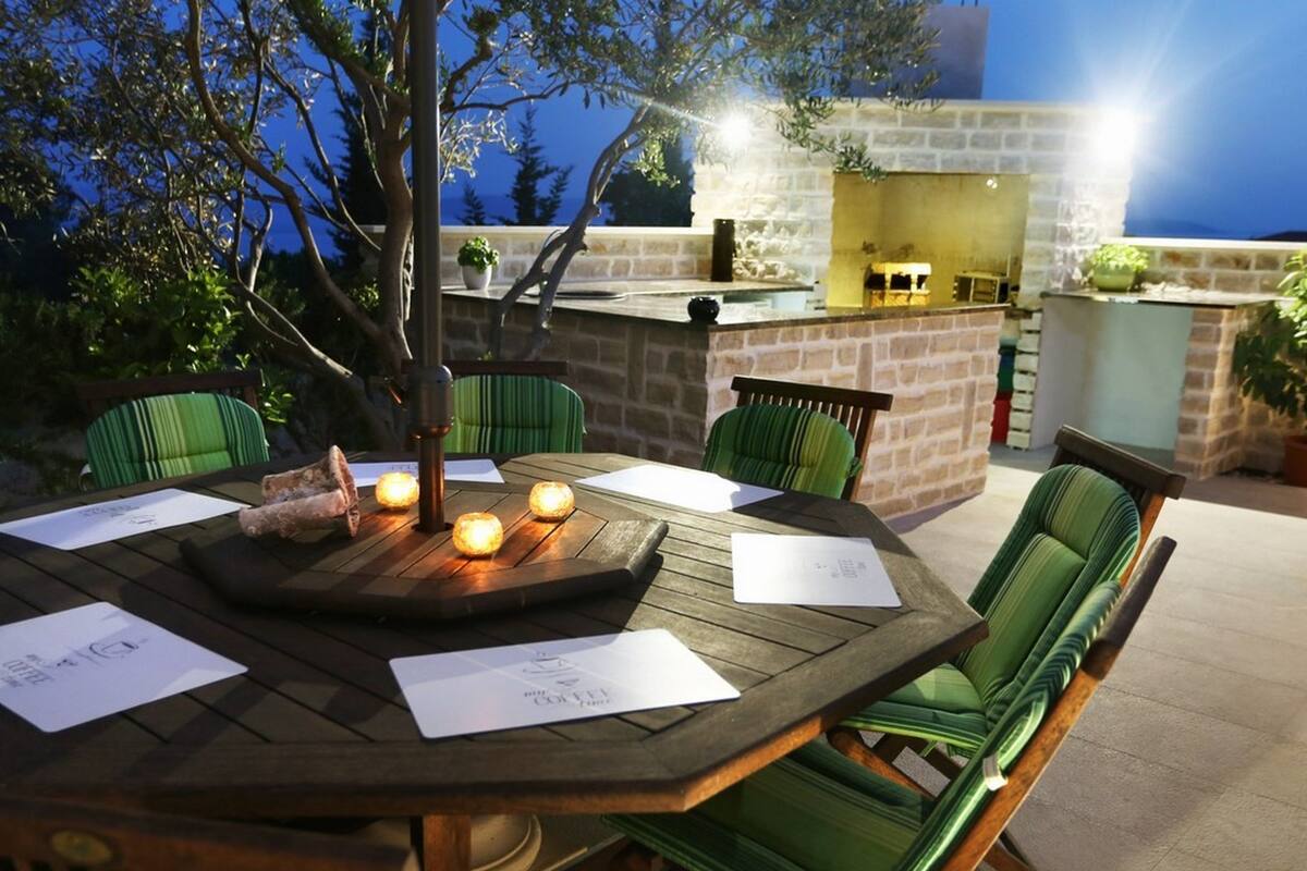 An outdoor dining area with a wooden table and chairs, a small tree, and a stone wall in the background.