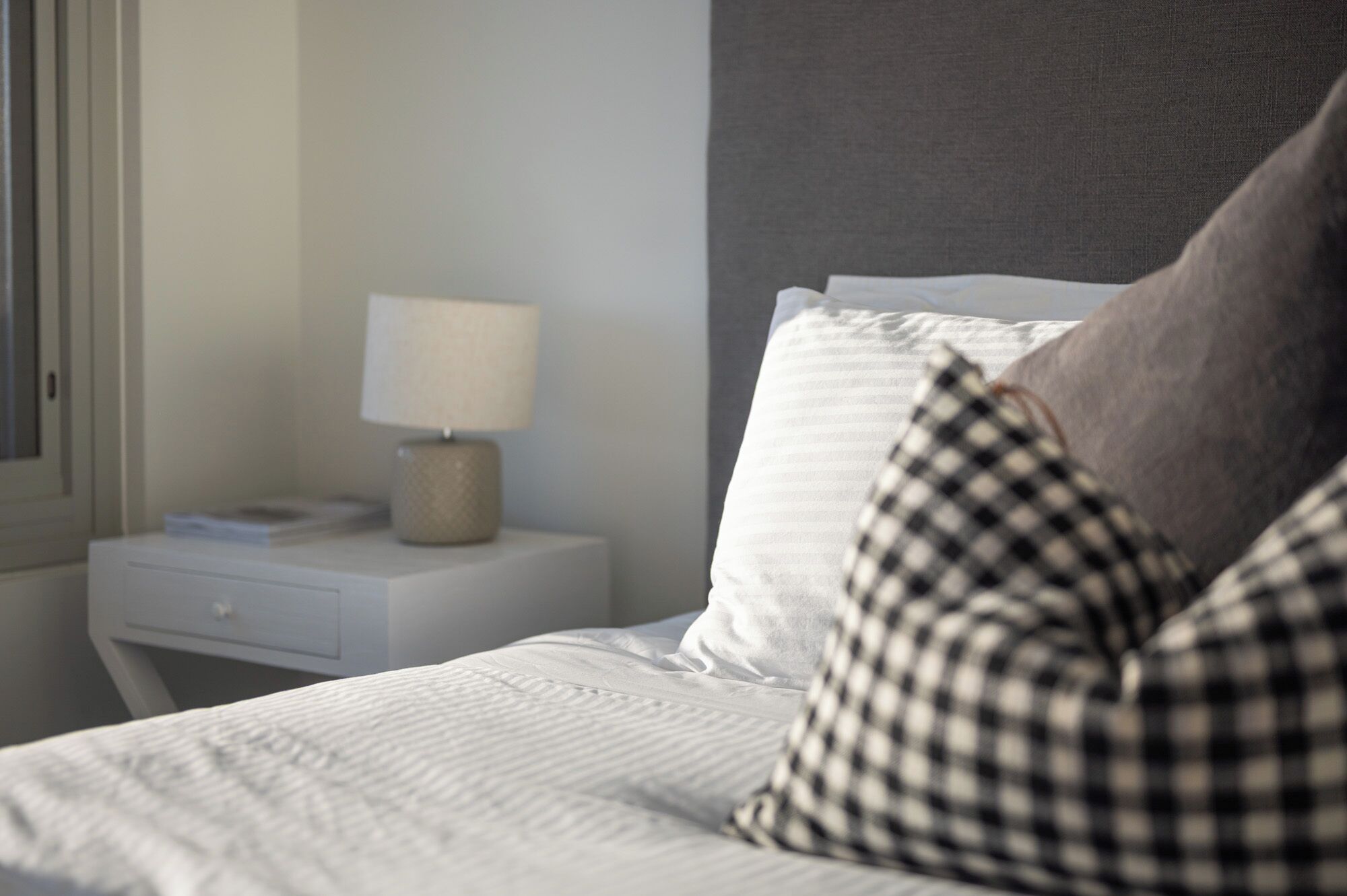 A neatly made bed with white bedding and a checkered pillow, a bedside lamp on a white nightstand, and a window with blinds.
