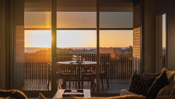 Ein Zimmer mit Blick auf den Sonnenuntergang über dem Meer, ein Esstisch mit Stühlen und eine Couch mit Kissen.