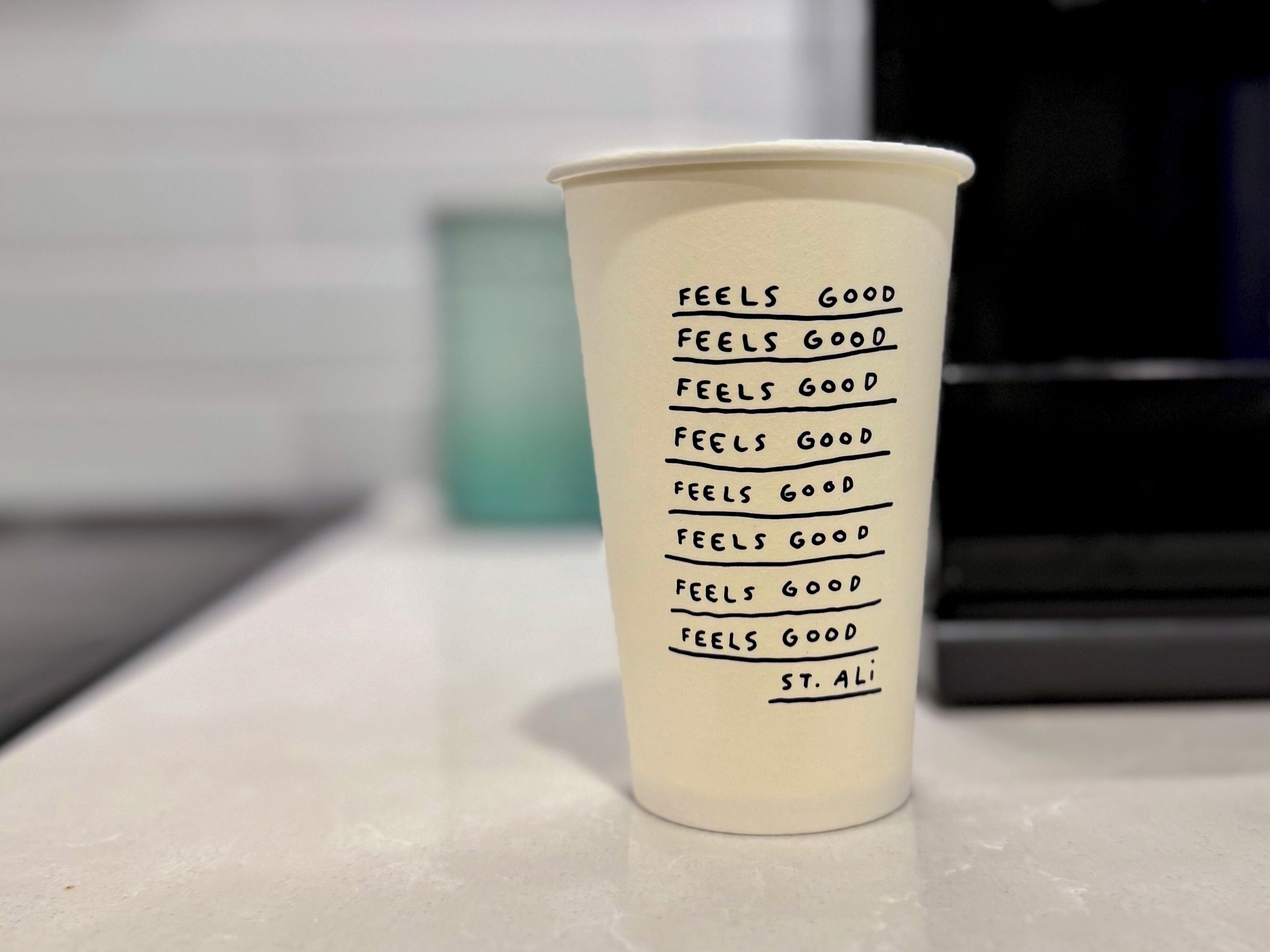 A white paper cup with the text "FEELS GOOD" repeated multiple times and "ST. ALI" at the bottom.