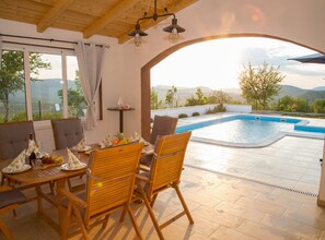 A poolside dining area with a view of mountains, a pool, and a table set for two with chairs. - Peaceful Escape With Mountain Views, Heated Pool and Modern Amenities in Radošić (Lecevica)