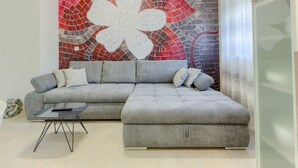 Ein Wohnzimmer mit einem grauen Ecksofa, einem kleinen schwarzen Couchtisch und einer großen, abstrakten Wand aus Mosaiksteinen.