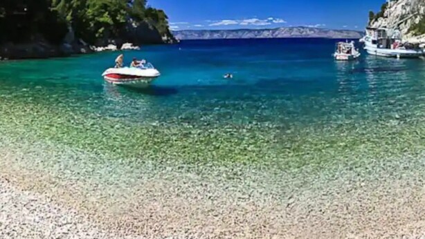 Une mer d’un bleu turquoise limpide, parsemée de bateaux, bordée de falaises rocheuses et d’une plage de galets.