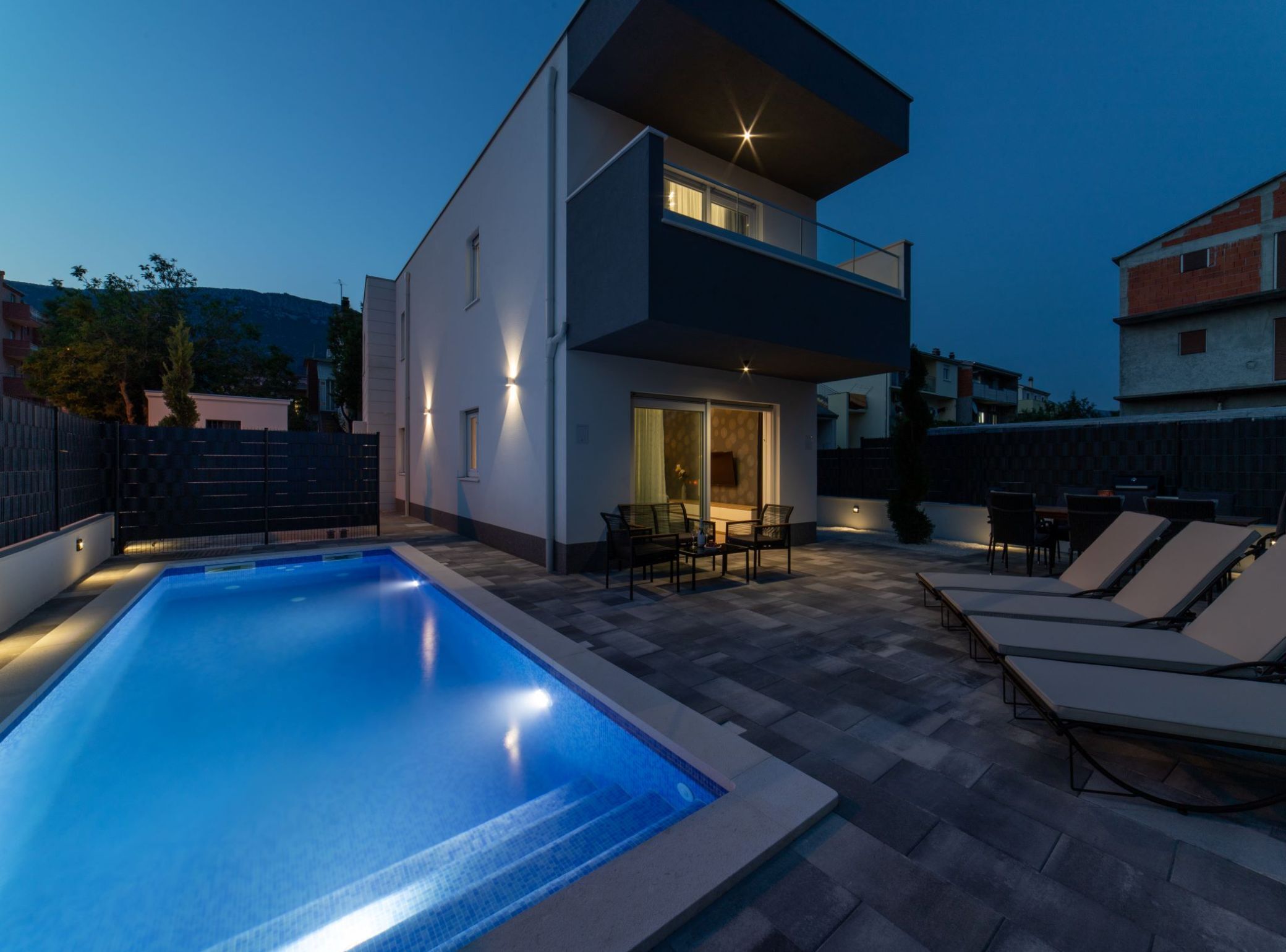 A modern two-story house with a swimming pool, outdoor seating, and a well-lit balcony.