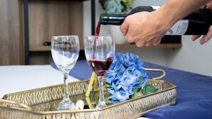 Eine Person gießt roten Wein in ein Glas auf einem Tablett mit goldfarbigem Henkel. Im Hintergrund befindet sich eine blaue Blumenkomposition und ein Regal mit gefalteten Handtüchern.