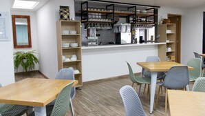 Ein modernes Café mit Holztischen und grünen Stühlen, einer Theke mit Glasware und Regalen mit Geschirr und Tassen.