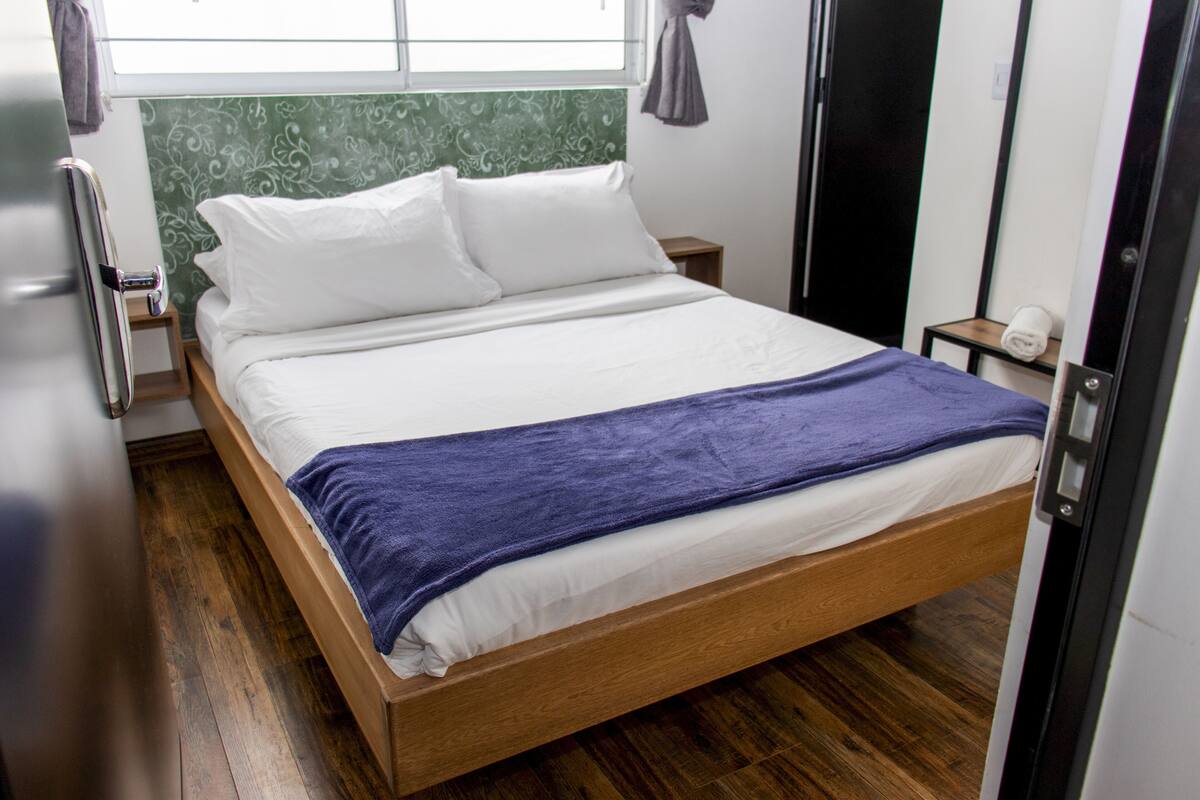 A neatly made bed with white linens and a blue blanket, a wooden headboard, a small bedside table with a lamp, and a window with curtains.