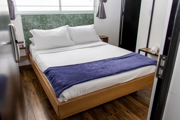 A neatly made bed with white linens and a blue blanket, a wooden headboard, a small bedside table with a lamp, and a window with curtains.