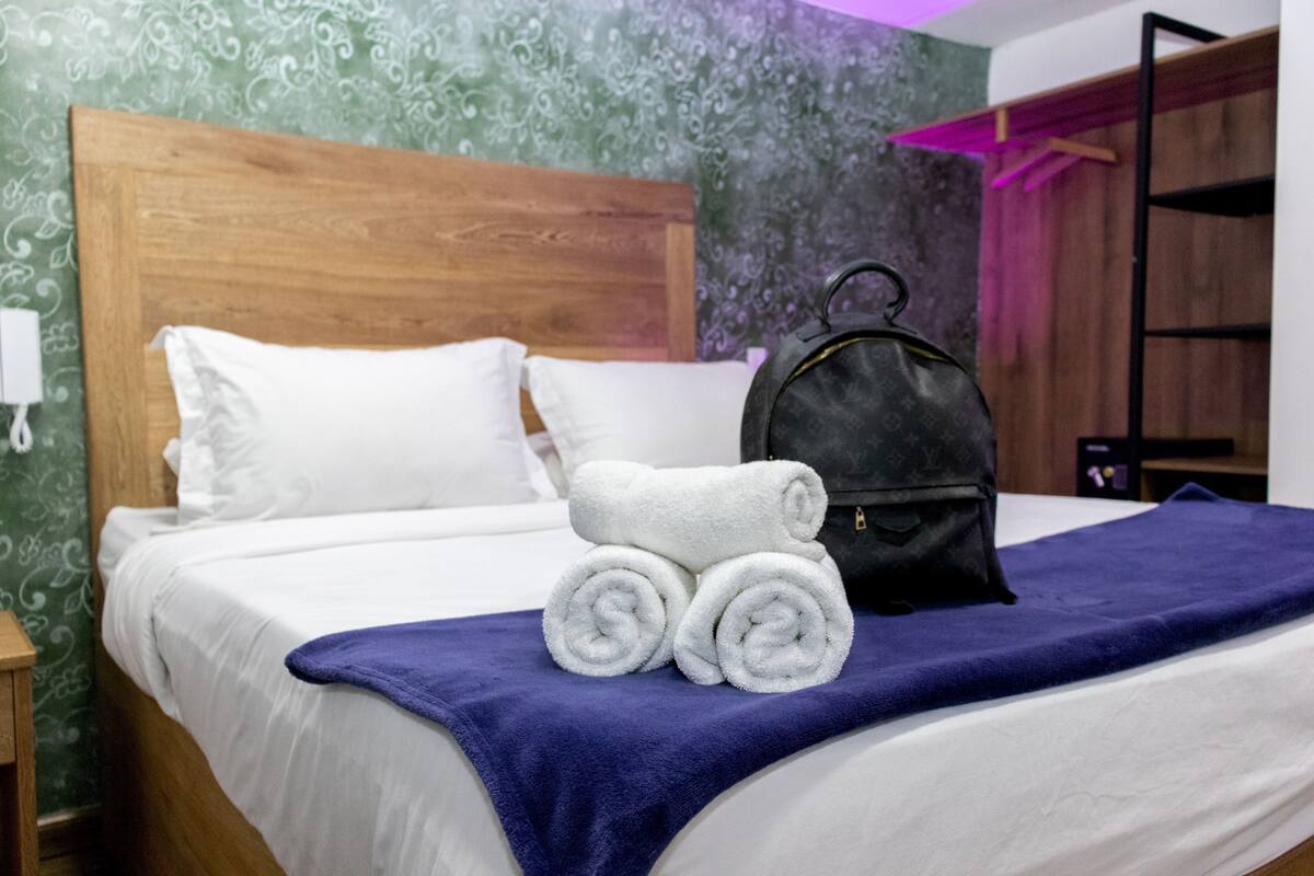 A neatly made bed with white linens, a blue blanket, and rolled towels. A black bag is placed on the bed.